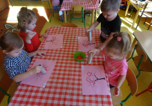 Czwórka dzieci stempluje rolkami papierowymi w kształcie serca koronę drzewa.
